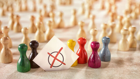 Symbolbild Wahl: Spielfiguren in verschiedenen Farben versammeln sich um einen Zettel, auf dem ein rotes Kreuz auf einem Kreis zu sehen ist.