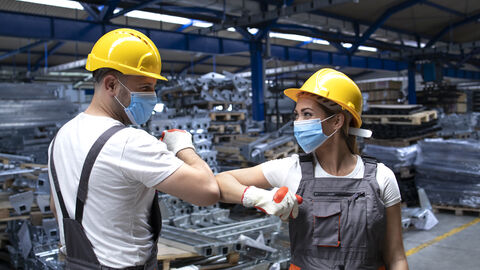 Ein Arbeiter und eine Arbeiterin in einer Industriehalle mit Masken, gelben Sicherheitshelmen und Arbeitslatzhosen begrüßen sich coronakonform per Ellbogencheck.