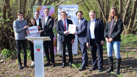 Staatssekretär Stefan Sauer (Mitte) steht gemeinsam mit den Beteiligten vor Bäumen und übergibt den Förderbescheid.