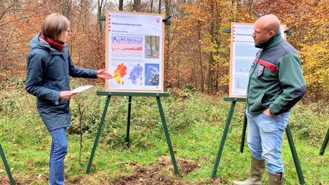Eine Frau zeigt einem Mann auf einem Plakat etwas, das auf einer Staffelei steht. Sie befinden sich in einem Wald