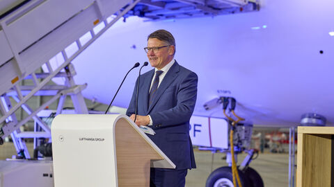 Ein Mann steht an einem Rednerpult. Im Hintergrund ist das Flugzeug.