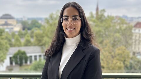 Frau mit dunklen Haaren mit weißem Rollkragenpullover und dunklem Blazer lächelnd vor Stadtpanorama
