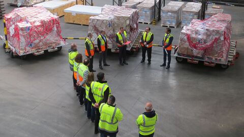 Mehrere Personen in gelben Warnwesten stehen zwischen Paletten mit Hilfsgütern am Flughafen.