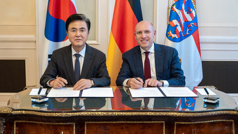 Zwei Männer sitzen nebeneinander an einem Tisch und sind gerade dabei jeweils ein Dokument zu unterzeichnen.