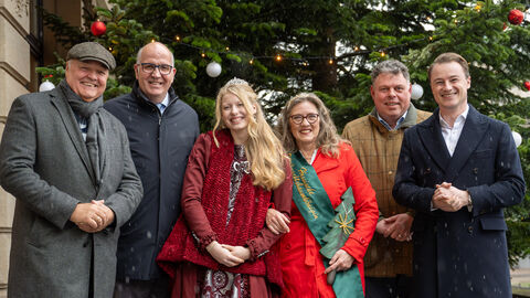Eine Gruppe Personen steht vor einem Nadelbaum zum Gruppenfoto aufgestellt. Alle lächeln in die Kamera.